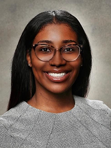Professional headshot of a person with shoulder-length dark hair, wearing round eyeglasses and a black-and-white patterned top.